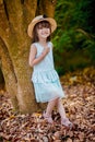 Beautiful little girl in a hat and white dress stands near a tree on a beautiful autumn warm day. Royalty Free Stock Photo