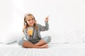 Beautiful little girl in headphones holding glass of orange juice, pointing with finger, looking at camera while sitting in bed Royalty Free Stock Photo