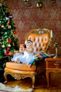 Beautiful little girl sits on a chair near a festive Christmas tree Royalty Free Stock Photo