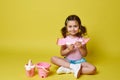 A beautiful little girl sits in a lotus position near pink beach toys on a yellow background, holds a shovel and rake, smiles Royalty Free Stock Photo