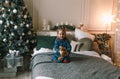 a beautiful little girl is sitting on the bed hugging a plush toy near a decorated Christmas tree Royalty Free Stock Photo