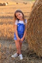 Beautiful little girl stands near a haystack in a summer field