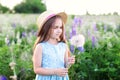 Beautiful little girl in a straw hat and dress holds a dandelion in a flowering field. Nature concept. A smiling girl is playing o