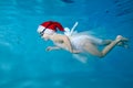 A beautiful little girl, wearing a Santa hat and a white dress, swims under the water in the pool and smiles. Portrait Royalty Free Stock Photo