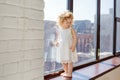 A beautiful little girl in a white dress stands by a large window in the sunlight Royalty Free Stock Photo