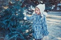 The beautiful little girl in winter wood. The girl is dressed in a gray fur coat. She is holding a white Christmas ball