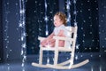 Beautiful little toddler girl in a white rocking chair in a dark room with Christmas lights Royalty Free Stock Photo