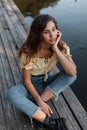 Beautiful lonely young curly girl in a yellow top with blue jeans sits on a wooden pier near the water Royalty Free Stock Photo