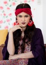 A beautiful long black hair lady with a red turban and purple blanket is sitting with elegant gesture on a big dark brown sofa in Royalty Free Stock Photo