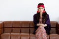 Beautiful long black hair lady with a red turban and purple blanket is sitting with her leg crossed on a big dark brown sofa in Royalty Free Stock Photo