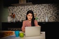 Beautiful long-haired Asian woman sits on her laptop in the kitchen at home where it`s convenient to eat during work.