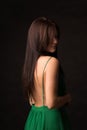 A beautiful long-haired brunette in a green dress with a bare back stands on a dark background. Romantic, pensive and sexy Royalty Free Stock Photo