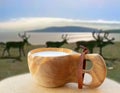 A handmade Nordic wooden cup with reindeer milk. In the background are reindeers.