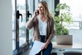 Beautiful mature business woman talking on mobile phone while holding papers looking through the window in the office Royalty Free Stock Photo