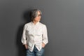 Beautiful mature lady in glasses stands hands in pockets looking at side. Elegant Gray-haired woman in whiite shirt on