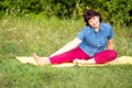 A beautiful mature woman sits on a sports mat and practices yoga in a forest park on a summer day. Royalty Free Stock Photo