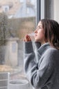 Beautiful melancholic teenager girl in warm grey sweater looking outside through raindrops on wet window at sky at spring rainy da