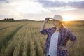 A beautiful middle-aged farmer woman in a straw hat and a plaid shirt stands in a field of golden ripening wheat during the daytim