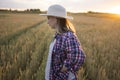 A beautiful middle-aged farmer woman in a straw hat and a plaid shirt stands in a field of golden ripening wheat during the daytim