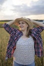 A beautiful middle-aged farmer woman in a straw hat and a plaid shirt stands in a field of golden ripening wheat during the daytim