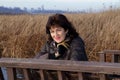 A beautiful middle-aged woman in a brown jacket sits and smiles against the backdrop of thickets of reeds by the river