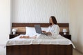 Beautiful middle aged woman dressed in white bathrobe sitting on bed and working with laptop in hotel room Royalty Free Stock Photo