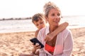 Beautiful middle aged woman with her son using phone in relaxing sitting at the beach Royalty Free Stock Photo