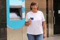 Beautiful middle aged woman is showing credit card near bank cash machine outdoors. Mature female withdrawing money with Royalty Free Stock Photo