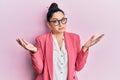 Beautiful middle eastern woman wearing business jacket and glasses clueless and confused expression with arms and hands raised