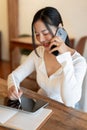 A beautiful Asian woman is talking on the phone while using her tablet, working remotely at a cafe Royalty Free Stock Photo