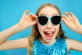 Beautiful mischievous Caucasian little girl in sunglasses close-up on a blue background. The girl is dressed in a