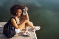 Beautiful mixed race female teenager sits on wooden pier and drinks cocktail in plactic glass