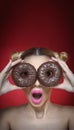 Beautiful model girl with bright pink lipstick holding two chocolate donuts. Funny joyful woman with sweets on dark red background