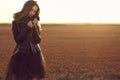 Beautiful model with long hair wearing black veiling dress and stylish leather jacket standing in the deserted field at sunset