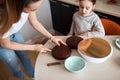 Beautiful mom and daughter are preparing birthday cake together, mom is cutting sponge cake using knife in kitchen. Royalty Free Stock Photo