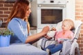 Beautiful mother in blue dress feeding with spoon her cute baby girl witch sits in high chair. Royalty Free Stock Photo