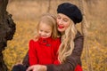 Beautiful mother in a long dress is swinging on a hinged swing little daughter in a red coat in an autumn garden near an old gnarl