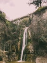 Beautiful mountain waterfall with a falling stream of water flowing from under a bridge on a cliff overgrown with trees and plants Royalty Free Stock Photo