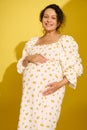 Beautiful pregnant woman in sundress, gently caresses her belly, smiles looking at camera, isolated on yellow background Royalty Free Stock Photo