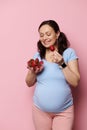 Beautiful pregnant woman holding bowl of ripe organic fresh strawberries, having healthy lunch, isolated pink background Royalty Free Stock Photo