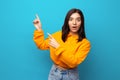 Beautiful multiethnicity woman in orange trendy shirt happily surprised expression pointing up against blue background