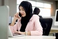 Beautiful Muslim Asian woman officer with hijab, talking with customer by mobile phone while working on laptop computer at office Royalty Free Stock Photo