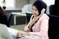 Beautiful Muslim Asian woman officer with hijab, talking on landline phone with customer while working on laptop computer at Royalty Free Stock Photo