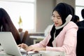 Beautiful Muslim Asian woman officer with hijab, talking on landline phone with customer while working on laptop computer at Royalty Free Stock Photo