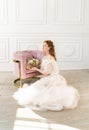 Beautiful natural redhead girl bride, with nude makeup, wearing a white dress, holds a wedding bouquet in her hands and sits on Royalty Free Stock Photo