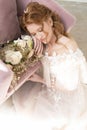 Beautiful natural redhead girl bride, with nude makeup, wearing a white dress, holds a wedding bouquet in her hands and sits on Royalty Free Stock Photo