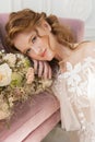 Beautiful natural redhead girl bride, with nude makeup, wearing a white dress, holds a wedding bouquet in her hands and sits on Royalty Free Stock Photo