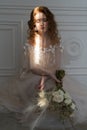 Beautiful natural redhead girl bride, with nude makeup, wearing a white dress, holds a wedding bouquet and sits on the floor in Royalty Free Stock Photo