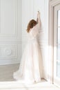 Beautiful natural redhead girl bride, with nude makeup, wearing a white dress,  stands turning her face to a white wall, holding a Royalty Free Stock Photo