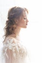 Beautiful natural redhead girl bride, with nude makeup, wearing a white dress, stands at the window next to a transparent curtain Royalty Free Stock Photo